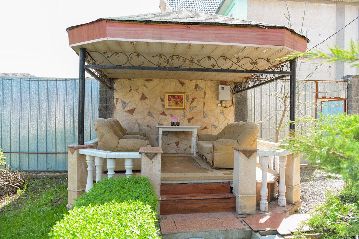 Беседка на территории: мягкие кресла и столик под навесом, каменная балюстрада, кустарник вокруг