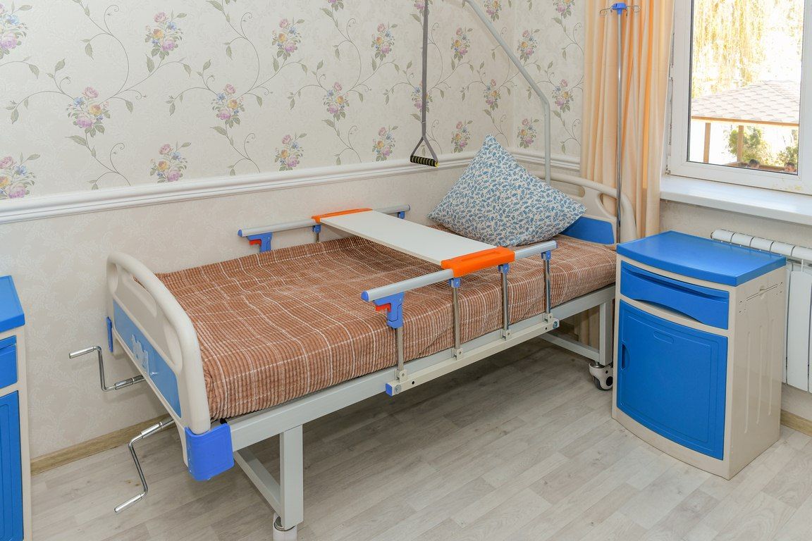 Функциональная кровать с подъёмным механизмом и прикроватным столиком, тумбочка и окно с занавеской рядом